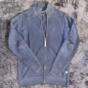 Mörkblå stickad kofta med dragkedja Från Indicode - Säljer en mörkblå stickad kofta från Indicode med grov ribbad krage, muddar och nederkant. Koftan har hel dragkedja framtill och en liten logotyp vid höften. Perfekt att slänga på över en t-shirt för en chill look.