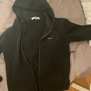 Svart hoodie från Comme des Garçons Play - Svart hoodie från Comme des Garçons Play med dragkedja och huva. Ikoniskt hjärta med ögon på bröstet och stort hjärta med ögon på. S/M