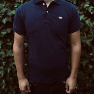 Mörkblå Lacoste pikétröja - Snygg mörkblå pikétröja från Lacoste med klassisk krage, två knappar och den ikoniska krokodilloggan på bröstet. Tillverkad i bomull för en skön känsla och perfekt passform. Enkel och stilren design som funkar till det mesta.                                                                                                                                                                              Nypris: 1350kr GORILLAPRIZE:620kr            Skick:10/10 oöppnad         KVITTO MEDKOMMER     
