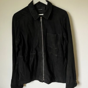 Svart Jonah mockajacka från J.Lindeberg - Stilren svart mockajacka från J.Lindeberg i getmocka. Jackan har klassisk krage, bröstficka och hel dragkedja framtill. Perfekt för dig som gillar minimalistisk och clean look. Passar till många olika outfits och ger en lyxig känsla. Modell: Jonah Zip Flat Suede - 1F14