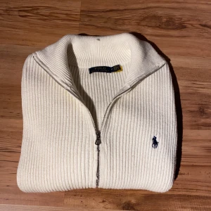 Vit ribbad kofta från Polo Ralph Lauren - Säljer en vit ribbad kofta från Polo Ralph Lauren med hel dragkedja och klassisk krage. Tröjan har lång ärm och den ikoniska blå Polo-loggan broderad på bröstet. Perfekt för dig som gillar stilrena och tidlösa plagg.