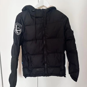 Svart pufferjacka från db - Svart pufferjacka från db med broderad patch på ärmen. Jackan har huva, dragkedja framtill och snörning vid halsen. Perfekt för kalla dagar och har en clean, sportig look som funkar till det mesta.
