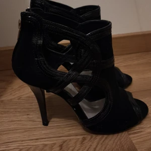 Svarta klackar från Bianco - Snygga svarta klackar från Bianco med höga klackar och öppen tå. Skorna har coola utskurna detaljer på sidorna och dragkedja bak. Materialmix av mocka och skinn ger en lyxig känsla. Perfekta för dig som vill sticka ut med en edgy look.