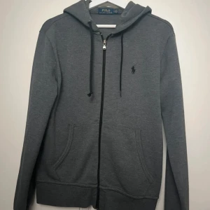 Grå zip hoodie från Polo Ralph Lauren - Snygg grå hoodie från Polo Ralph Lauren med dragkedja, huva och svarta snören. Klassisk logga broderad på bröstet och två stora fickor framtill. Perfekt för chill dagar eller när du vill ha en clean streetstyle.