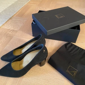 Stinaa J, modell ”Alice”, 6 cm klack - Stilrena svarta pumps från Stinaa.J i mjuk mocka med spetsig tå och stabil klack, 6 cm. Skorna har en diskret gulddetalj vid hälen och en klassisk siluett som passar till många looks. Levereras med originalkartong och dustbag. Väldigt sparsamt använda. Nypris ca 4.500 kr.