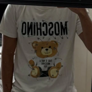 Vit Moschino t-shirt med nalletryck - Säljer en vit t-shirt från Moschino med stort tryck av en gullig nalle och säkerhetsnålar på framsidan. T-shirten har rund hals och korta ärmar. Materialet är mjuk bomull och passformen är normal. Perfekt statement-plagg för dig som gillar coola designer.