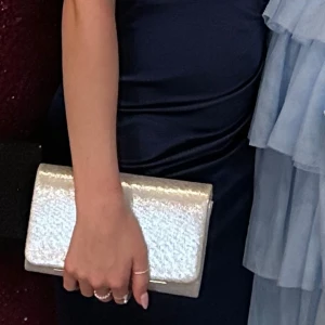 Silver kuvertväska i syntetmaterial - Säljer silvrig kuvertväska. Köpte begagnad och sedan använt den en gång till balen så i fint skick. Finns kjeda till oxå