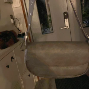 Grå axelväska i skinnlook - Snygg grå axelväska med rundad form och bred axelrem med metallringar. Väskan har magnetlås och ett rymligt huvudfack. Perfekt för dig som vill ha en stilren och enkel väska till vardags. Materialet har en matt skinnkänsla och insidan är fodrad i mörkt tyg.