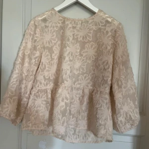 Beige blommig blus med spetsdetaljer - Romantisk beige blus med stora blommiga spetsdetaljer och lätt transparent tyg. Blusen har trekvartsärmar och en lös passform som ger en luftig känsla. Perfekt för dig som gillar en feminin och trendig stil.