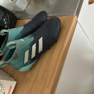 Adidas fotbollsskor i blått och turkos - Säljer ett par Adidas fotbollsskor i snygg blå och turkos färg med klassiska vita ränder på sidan. Skorna har en strumpliknande passform utan snörning och dobbar för gräsplan. Perfekta för dig som vill ha stil och funktion på planen.
