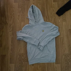 Ljusgrå hoodie från Lyle & Scott - Säljer en ljusgrå hoodie från Lyle & Scott med klassisk gul logga på bröstet. Tröjan har huva med snörning och en stor magficka. Perfekt för dig som gillar en enkel och stilren look.