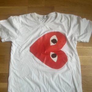 Vit Comme des Garçons Play t-shirt - Säljer en vit t-shirt från Comme des Garçons Play i storlek L. T-shirten har ett stort rött hjärta med ögon tryckt på framsidan. Klassisk rund hals och korta ärmar. Materialet är mjuk bomull, perfekt för en chill och stilren look. Använd en gång 