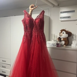 Röd glittrig balklänning Jolie Bride - Säljer en magisk röd balklänning från Jolie Bride med glittriga paljetter upptill och v-ringad urringning. Klänningen har en lång, voluminös kjol i tyll och är ärmlös för en elegant look. Perfekt för bal eller festliga tillfällen!