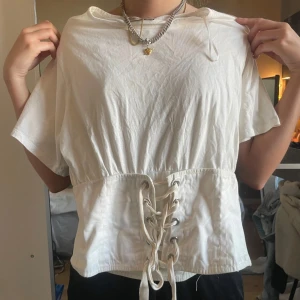 Vit kortärmad topp med snörning cool fest topp - Cool fest topp Säljer en stilren vit topp med korta ärmar och snörning i midjan. Perfekt för en avslappnad look med en twist. Toppen har en lös passform och är tillverkad i ett mjukt material. Notera att dragkedjan bara funkar ibland så brukar sätta fast den med en säkerhets nål 