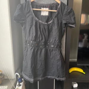 Unik blus från Esprit - Svart blus från Esprit med korta ärmar och rund halsringning. Blusen har dekorativa paljetter och pärlor längs bysten och midjan, vilket ger en snygg detalj. Tillverkad i mjukt bomullsmaterial och passar perfekt till många olika outfits.