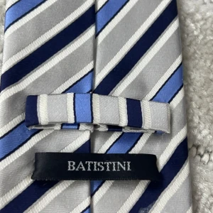 Randigt slips från Batistini - Säljer en snygg slips från Batistini med diagonala ränder i blått, silver, vitt och svart. Slipsen har en klassisk form och är tillverkad i ett glansigt material som ger en stilren look. Perfekt för att lyfta din outfit.