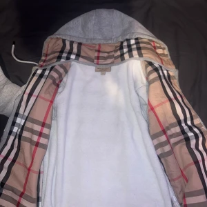 Grå burberry hoodiejacka med Burberry-mönstrat  - Grå zip-hoodie med klassiskt Burberry-rutigt foder i huva och insida. Jackan har dragsko i huvan och dragkedja framtill. Perfekt för en stilren och avslappnad look.