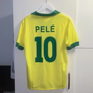 Brasilien fotbollströja Pelé 10 - Klassisk gul Brasilien fotbollströja med gröna detaljer, tryck 'BRASIL' och fem stjärnor på framsidan. Baksidan har grönt Pelé-tryck och nummer 10. Kortärmad modell med grön krage och ärmslut. Perfekt för fans av legendariska spelare.