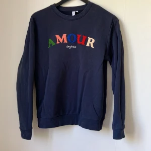 Mörkblå sweatshirt med broderad text från & Other Stories - Mörkblå sweatshirt från & Other Stories med färgglad broderad text 'AMOUR' på bröstet. Rund halsringning och långa ärmar. Perfekt för en avslappnad och trendig look.