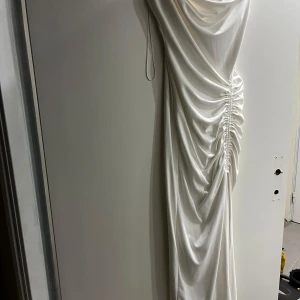 Vit långklänning med drapering från Zara - Elegant vit långklänning från Zara med snygg drapering och rynkade detaljer längs sidan. Klänningen har smala axelband och en mjuk, följsam passform som ger en lyxig känsla. Perfekt för speciella tillfällen när du vill sticka ut.
