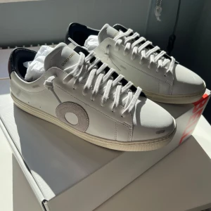 Oliver Cabells - Säljer ett par helt nya fräscha vita sneakers från Oliver Cabell med svart häl och grå cirkeldetalj på sidan. Skorna har snörning och platt sula. Säljer pga att jag köpte fel storlek. Skriv vid mer frågor och funderingar!