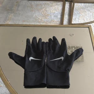 Svarta träningshandskar från Nike - Svarta, tunna träningshandskar från Nike med vit logga på ovansidan. Handskarna har en smidig passform och är perfekta för utomhusaktiviteter när det är lite kyligt. Materialet är stretchigt och följsamt.
