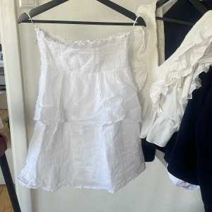 Vit volangklänning från Gina Tricot - Söt vit klänning från Gina Tricot med smock upptill och volangdetaljer. Axelbandslös modell med kort längd och lätt, luftig känsla. Perfekt för varma dagar och somriga tillfällen.