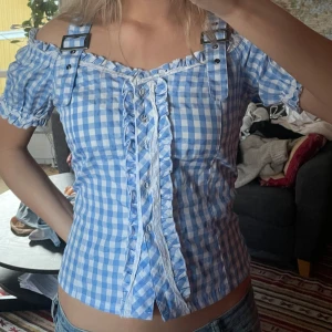 Blåvit rutig blus med volang och snörning - Supersöt blåvit rutig blus med offshoulder och puffärmar. Framtill finns snörning och volangdetaljer, samt dekorativa spännen på axlarna. Perfekt för en somrig look! från JANINA, sitter som strlk S på mig funkar typ vilken strlk som helst då det gpr attjustera båda midjan och axlarna