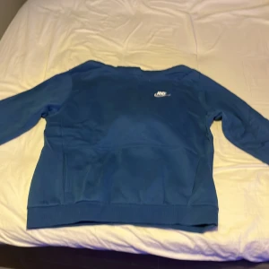Blå hoodie från Nike - Säljer en blå hoodie från Nike med broderad vit logga på bröstet. Tröjan har huva och ribbade muddar vid ärmslut och nederkant. Perfekt för en sportig och avslappnad stil.