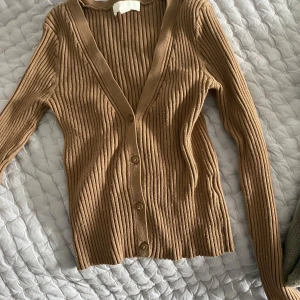 Beige ribbad kofta med v-ringning - Säljer en beige ribbad kofta med v-ringning och knappar framtill. Tröjan har lång ärm och är i ett mjukt, stretchigt material som sitter snyggt på kroppen. Perfekt att slänga över en topp eller bära som den är.