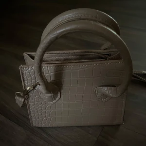 Beige mini handväska med krokodilpräglat mönster - Säljer en beige mini handväska med krokodilpräglat mönster och guldfärgade detaljer. Väskan har både handtag och axelrem samt dragkedja upptill. Perfekt för dig som vill ha en stilren accessoar med lyxig känsla.