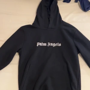 Svart hoodie från Palm Angels - Säljer en svart hoodie från Palm Angels i storlek M. Tröjan har vit Palm Angels-logga tryckt både fram och bak samt en klassisk huva och känguruficka. Perfekt för dig som gillar streetwear och vill ha något stilrent.