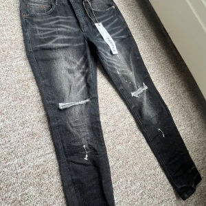 Svarta jeans från Purple Brand med slitningar - Säljer ett par svarta jeans från Purple Brand med slitningar på benen och lätt tvättad look. Jeansen har skinny passform, klassiska fem fickor och detaljer som dragkedja och knappar. Perfekta för dig som gillar en trendig och avslappnad stil.