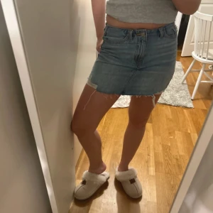 Blå jeanskjol med rå kant - Säljer en klassisk blå jeanskjol med rå, fransig kant nertill. Kjolen har fem fickor och stängs med dragkedja och knapp framtill. Perfekt för en avslappnad och trendig look.