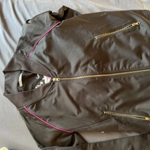 Svart bomberjacka med broderade rosor - Svart bomberjacka med dragkedja och rosa detaljer. På ryggen finns ett stort broderi med rosa rosor och dödskalle. Jackan har långa ärmar och två fickor med dragkedja framtill.