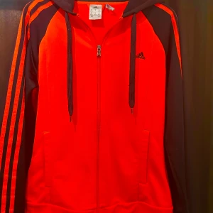 Röd och svart hoodie från Adidas - Säljer en snygg röd hoodie från Adidas med svarta detaljer och dragkedja framtill. Tröjan har huva med snören, svarta ärmar med röda ränder och två fickor. Perfekt för en sportig look eller chill vardagsstil.
