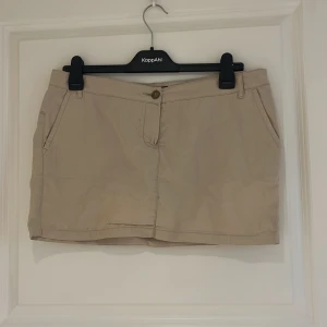 Beige kortkjol med fickor - Säljer en beige kortkjol med två fickor fram och dekorativa fickor bak. Kjolen har hällor för skärp och stängs med knapp och dragkedja. Perfekt för en enkel och stilren look.