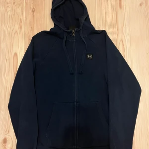 Mörkblå,grön och grå hoodie från Under Armour - Mörkblå hoodie med dragkedja från Under Armour. Tröjan har huva och två fickor framtill och en liten logotyp på bröstet. Perfekt för träning eller chill. Har också i GRÖN och GRÅ. 200 st
