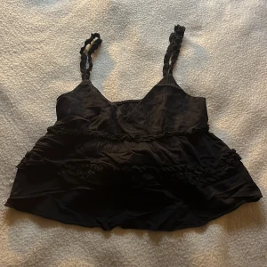 Svart volangig linnetopp - Säljer en svart linnetopp med volangdetaljer och smala axelband. Toppen har en luftig passform och är perfekt för varma dagar. Axelbanden är dekorerade med små volanger.