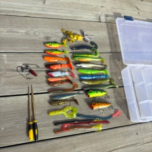 Fiskedrag med beteslåda - Säljer ett set med färgglada mjukbeten och jiggar i olika former och färger, perfekt för sportfiske. Medföljer en genomskinlig plastlåda med fack för enkel sortering samt två tänger och några extra krokar. Betena är i plast och gummi, samt 2 krokfilar. Bra skick på båda poppers och jiggarna.