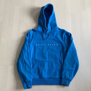 Blå hoodie från Daily Paper - Säljer en blå hoodie från Daily Paper med vit broderad logga på bröstet. Tröjan har huva med dragsko och en stor magficka framtill. Perfekt för en avslappnad och cool stil.