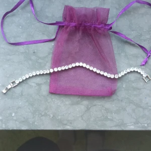 Silverfärgat armband med glittrande stenar🤩 - Ett elegant armband i silverfärg med en rad gnistrande, runda stenar. Armbandet har ett klassiskt lås och levereras med en lila organzapåse. Perfekt för att ge din outfit extra glans.