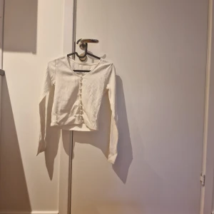 Vit ribbad långärmad topp med knappar - Säljer en vit ribbad långärmad topp med v-ringning och knappar framtill. Perfekt för att styla med jeans eller kjol. Enkel och stilren design som passar till många olika looks.