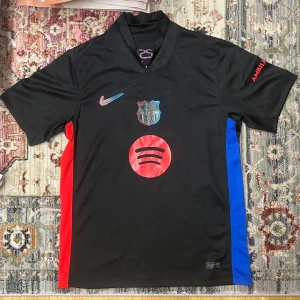 FC Barcelona svart borta matchtröja Lamine Yamal 19 - Säljer en svart FC Barcelona fotbollströja från Nike med Lamine Yamal 19 på ryggen. Tröjan har klubbmärke, Nike-logga och Spotify som sponsor framtill samt röda och blå paneler på sidorna. Tillverkad i Dri-fit material.