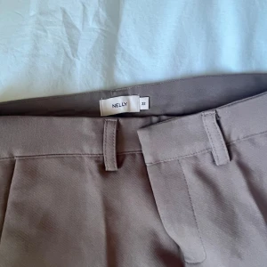 Beige kostymbyxor från Nelly - Snygga beige kostymbyxor från Nelly med klassisk passform och pressveck. Byxorna har hällor för bälte, snedställda fickor fram och dekorativa fickor bak. Perfekta för en stilren look.