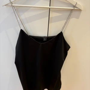 Svart linnetopp med kedjeband - Snygg svart linnetopp med tunna kedjeband som är glittriga som axelband. Perfekt för en trendig och stilren look. Toppens enkla design gör den lätt att matcha med olika outfits.