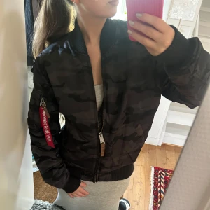 Alpha Industries bomberjacka - Säljer en svart bomberjacka med kamouflage-mönster från Alpha Industries. Jackan har dragkedja, ribbade muddar och orange foder. Klassisk röd tag på ärmen. Herrjacka i M, men passar även dam! Typ aldrig använd🥰