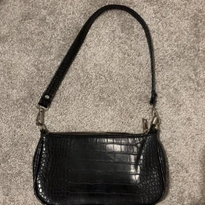 Svart axelväska i krokopräglat skinn - Snygg svart axelväska med krokodilpräglat mönster och dragkedja upptill. Väskan har en avtagbar axelrem och är perfekt för att bära det viktigaste. Stilren och trendig design som passar till många outfits.