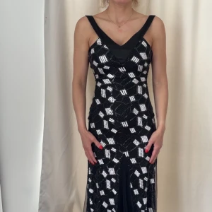 Svart långklänning med paljetter och snörning i ryggen - Säljer en svart långklänning med vita paljetter i geometriskt mönster. Klänningen har djup v-ringning, breda axelband och snörning i ryggen. Materialet är glittrigt och elegant, perfekt för  fest.