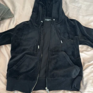 Svart velour hoodie från Juicy Couture - Säljer en svart hoodie i mjuk velour från Juicy Couture. Jackan har huva, dragkedja med J-detalj, två fickor fram och broderad JC-logga på bröstet. Perfekt för en avslappnad och trendig look.